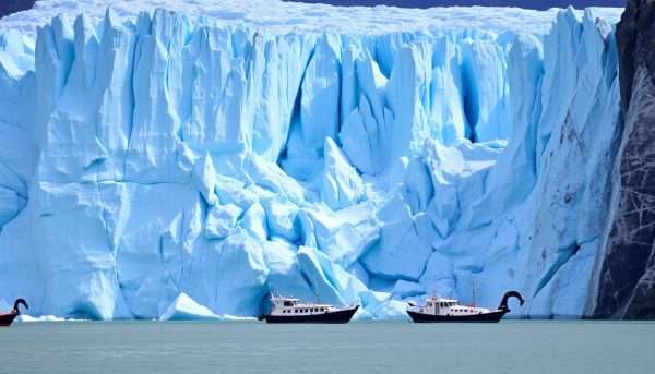 The massive blue ice wall of Perito Moreno Glacier with small boats in the foreground for scale The massive blue ice wall of Perito Moreno Glacier with small boats in the foreground for scale