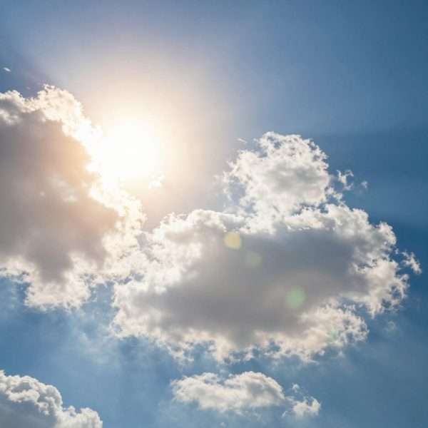 Bright sun with rays shining through fluffy white clouds on perfect blue sky day