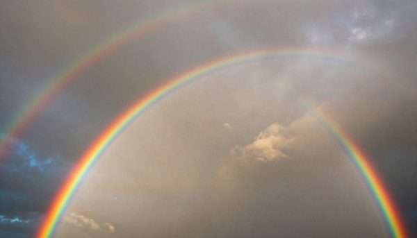 Vibrant double rainbow arcing across sky after rain with sun breaking through clouds